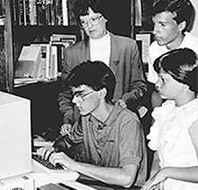 A young man uses a 1990s-era computer while others look on.