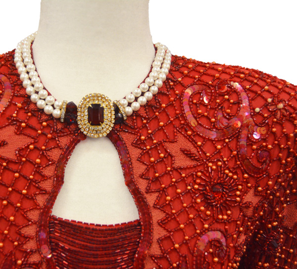 Close-up of the top of a red ball gown with intricate red beads of various sizes, and a necklace of pearls and red stones.