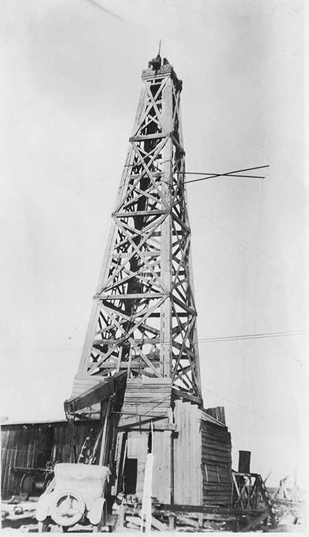 A historic photograph wooden oil well with a vehicle parked in front of it. 