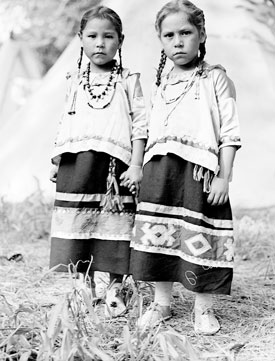 Two young girls with their hair in braids wearing beaded necklaces and traditional dance clothing. The girls are Ernestine Black and Betty Joe Buffalohead. 